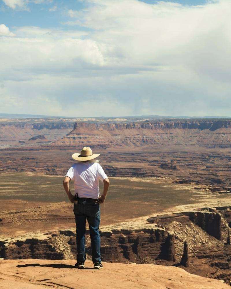 A man in a wide-brimmed hat stands at the edge of a canyon, looking out over miles of red rock country