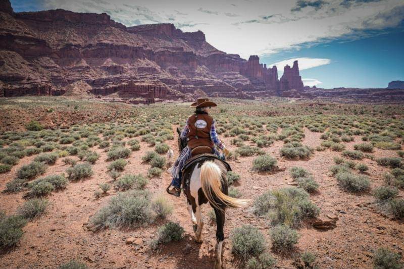 A rider on horseback crossing sage flats toward towering red cliffs
